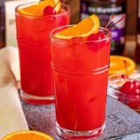 Square image close up of Alabama Slammer on metal tray garnished with orange and cherry.